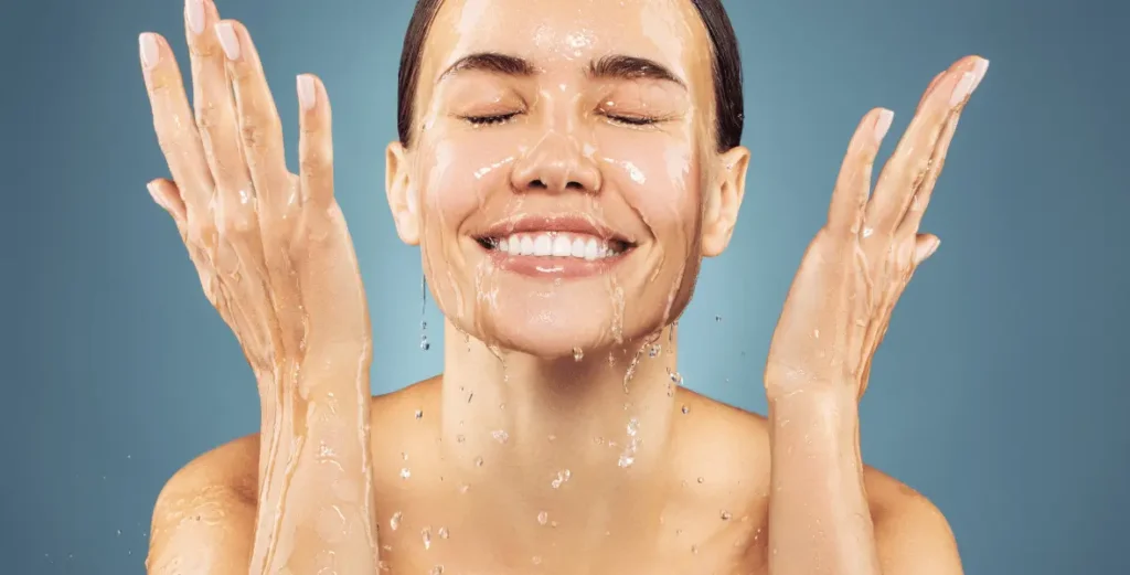 A woman splashing her face with water as if she is cleansing her skin, for an article about cleansing skin properly
